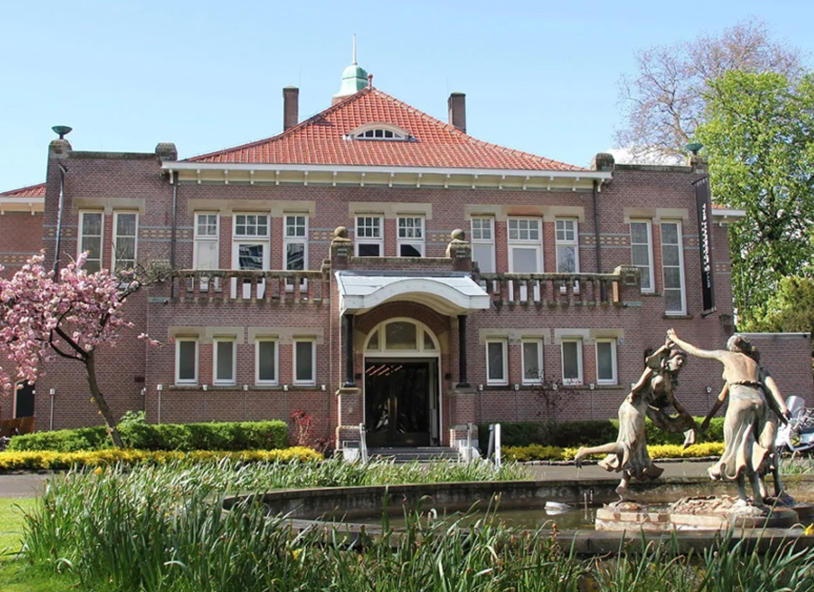 Open, sunlit park in Rotterdam with trees and green grass.